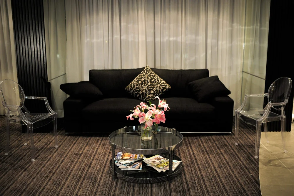 You Day Spa relaxation lounge — black sofa and ghost chairs before sheer curtains over the Melbourne skyline